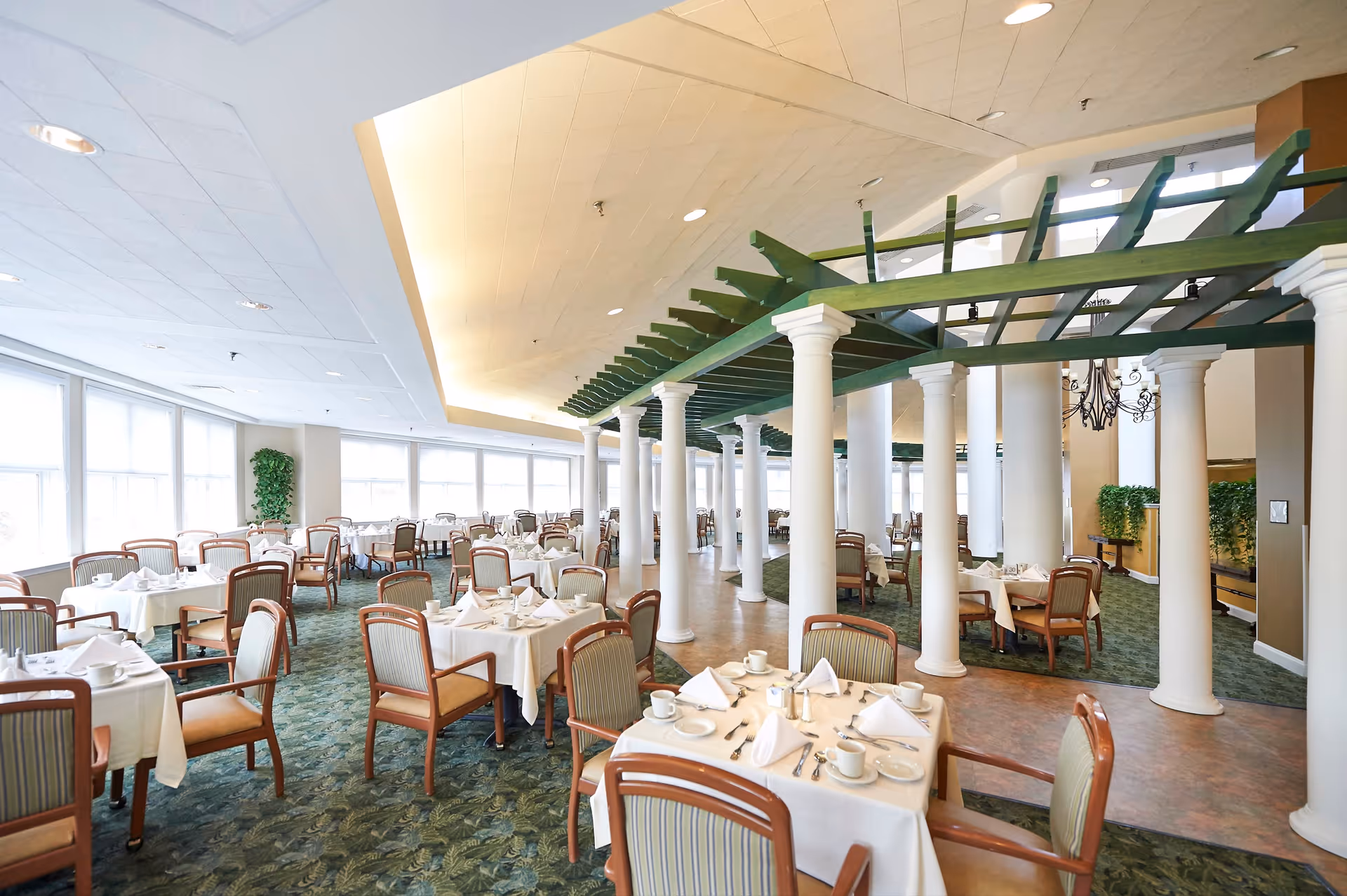 A spacious dining room with multiple tables covered in white tablecloths, each set with white napkins, cups, and silverware. The room features green carpet flooring, large windows letting in natural light, white columns supporting a green pergola structure, and beige walls with some greenery decorations.