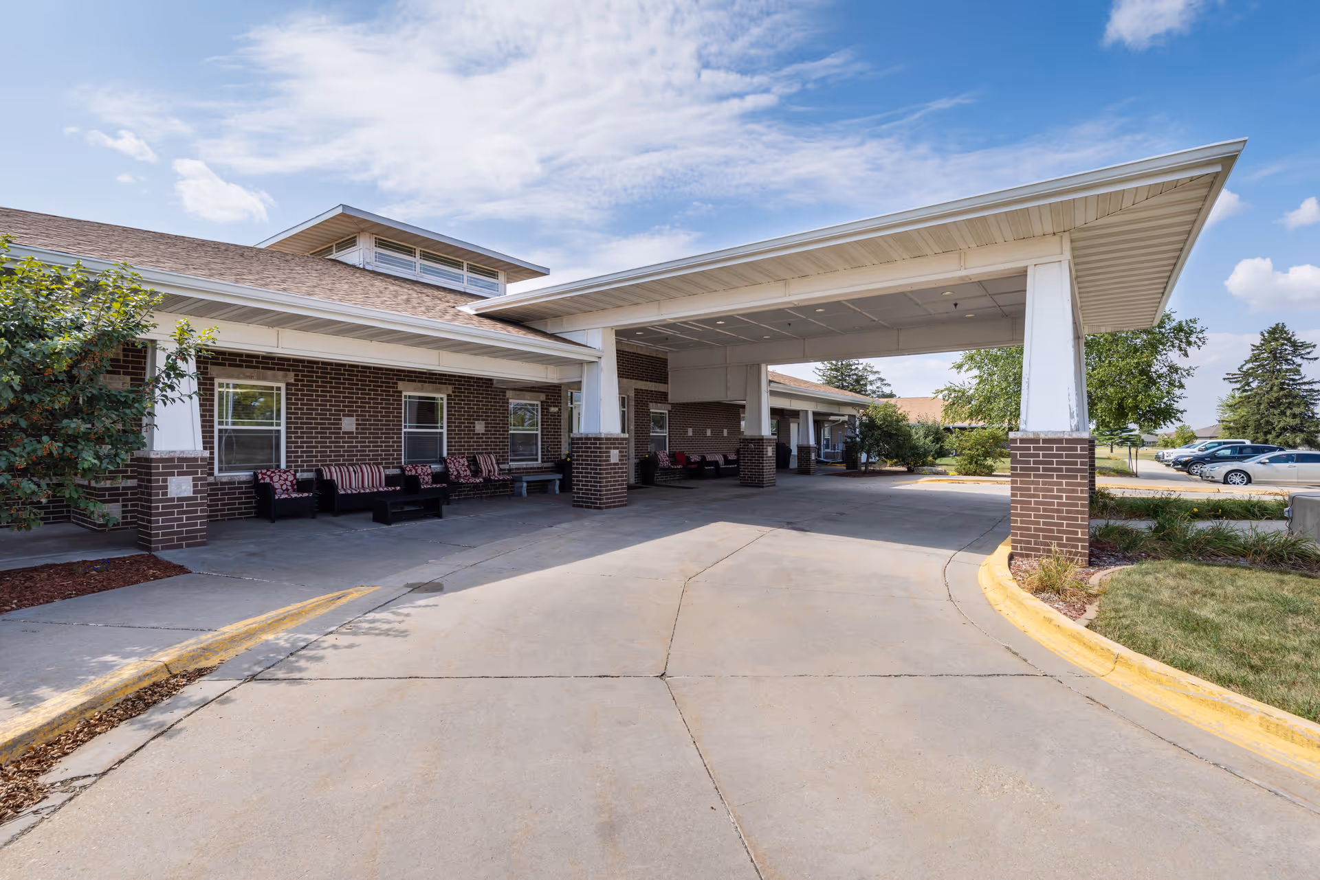 Covered drive-up entrance with outdoor seating at the front of a brick senior living facility under a partly cloudy sky.