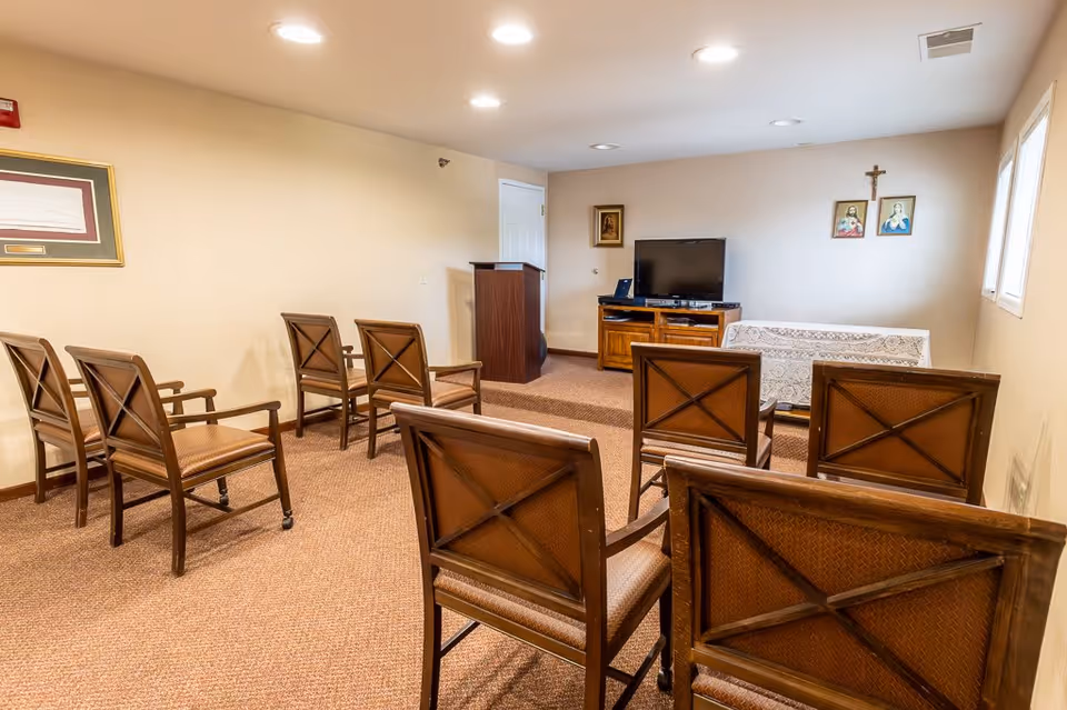 A small room with several wooden chairs arranged in rows facing a wooden podium and a TV on a wooden stand. The room has beige walls, carpeted floor, and religious pictures and a crucifix hanging on the wall behind the TV. There is a window on the right side letting in natural light.