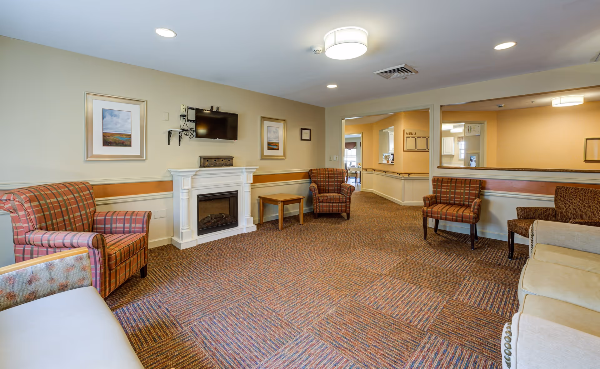 A cozy living room area with patterned armchairs and a beige sofa arranged around a white electric fireplace. Above the fireplace is a mounted flat-screen TV, and two framed landscape paintings hang on the wall. The room has a carpeted floor with a multicolored pattern, recessed ceiling lights, and a large mirror on one wall reflecting part of the adjacent hallway.