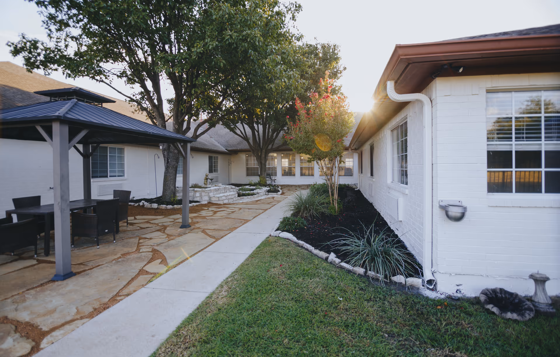 Outdoor courtyard of a white-brick memory care facility featuring a paved walkway, gazebo-style covered seating area, trees, and landscaped beds.