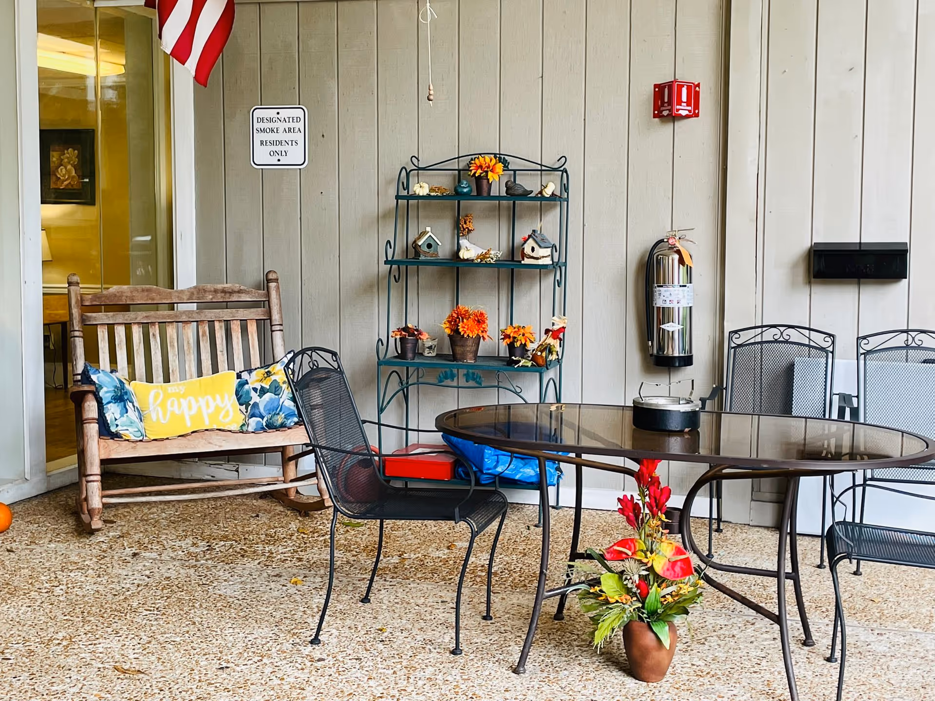 Outdoor patio area with a wooden rocking bench that has a yellow pillow with the word 'happy' on it, a glass-top metal table surrounded by metal chairs, a metal shelf with decorative items including small birdhouses and flower pots, a fire extinguisher mounted on the wall, and a sign that reads 'Designated Smoke Area Residents Only'.