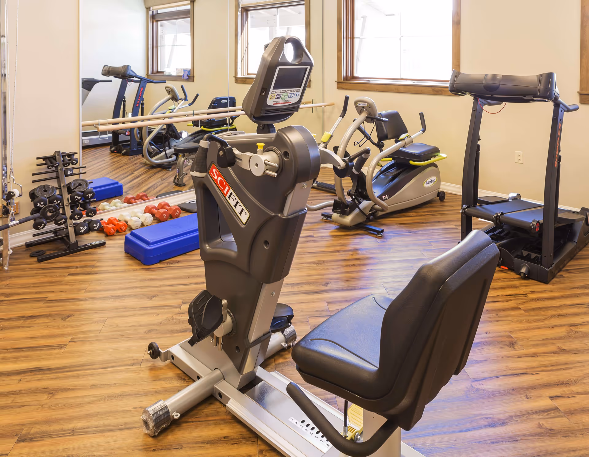A small fitness room with exercise equipment including a recumbent bike, treadmill, elliptical machine, and a set of dumbbells on a rack. The room has wooden flooring, two windows with wooden frames, and a large mirror on one wall.
