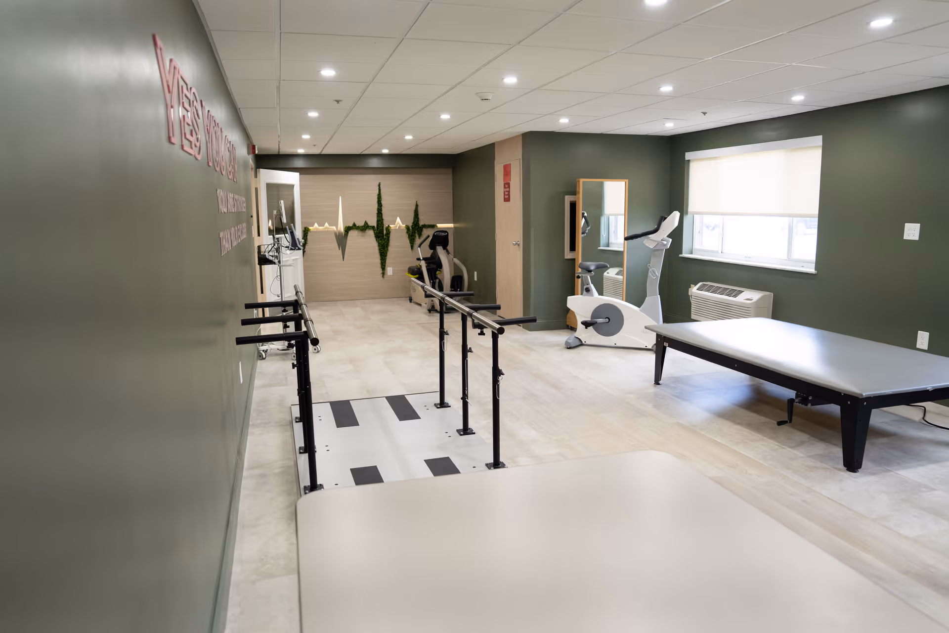 A rehabilitation room with green walls, featuring parallel bars for walking exercises, an exercise bike, a large mirror, and a therapy table. The room is well-lit with ceiling lights and has a window with a roller shade and an air conditioning unit below it. There is a decorative wall art piece resembling a heartbeat line with greenery.