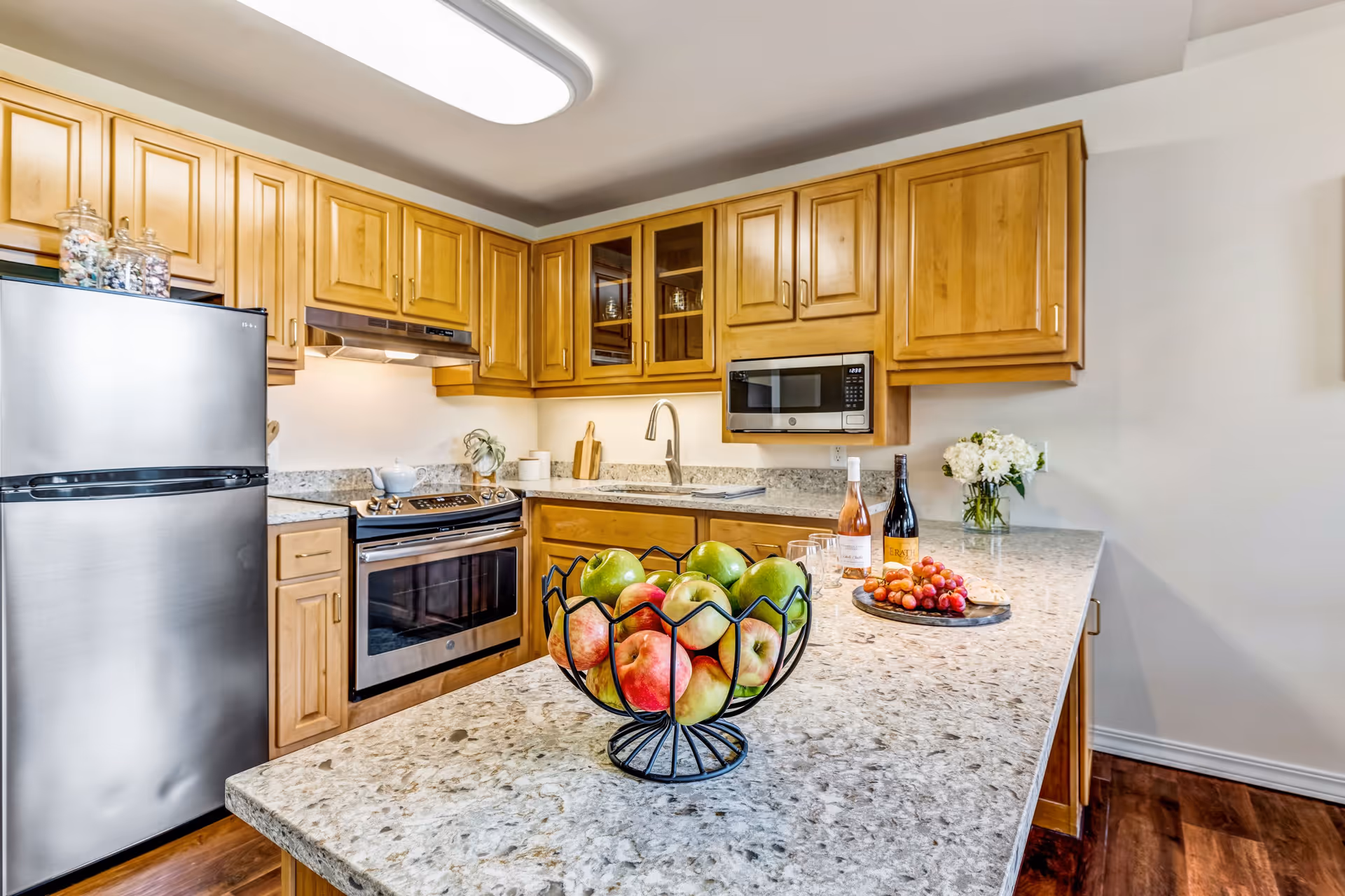 Modern kitchen with wooden cabinets, granite countertops, stainless steel refrigerator and oven, a microwave mounted above the counter, a bowl of apples on the island, and a tray with wine bottles and grapes on the counter.