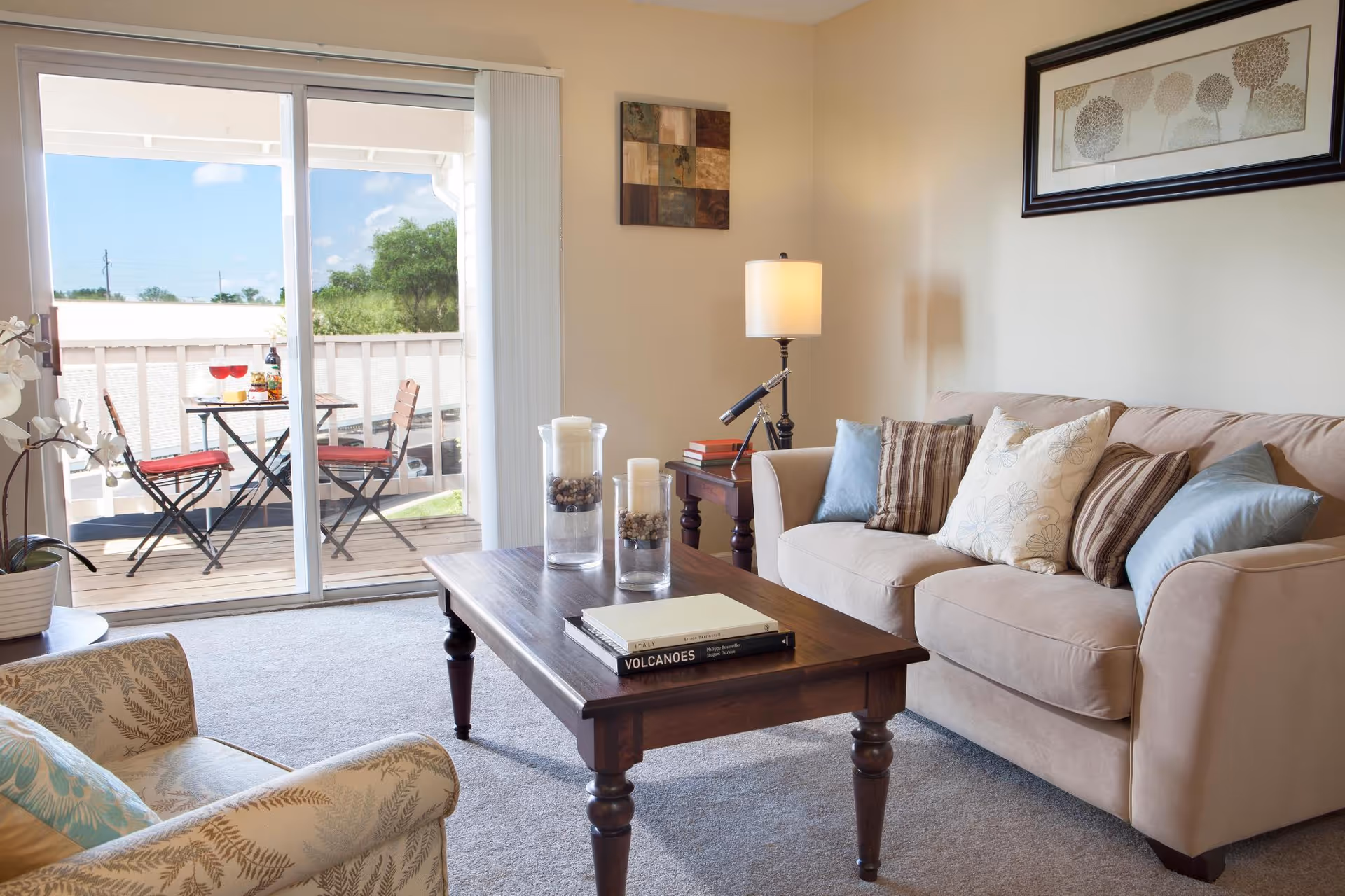 Sunlit living room with a beige sofa, armchair, wooden coffee table, lamp, and sliding glass door opening to a small balcony with a bistro table.