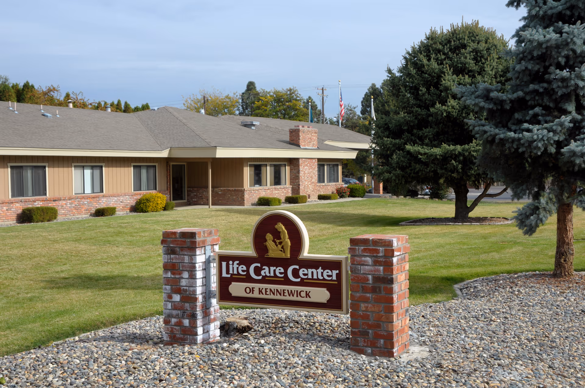 Exterior view of the Life Care Center of Kennewick building with a lawn, trees, and a sign in the foreground displaying the facility's name.