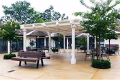 Outdoor patio area with benches, tables, and chairs under a white pergola structure surrounded by trees and greenery.