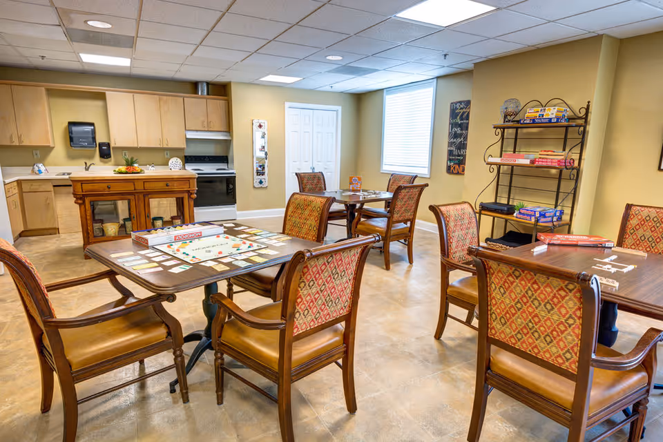 A bright room with several wooden tables and upholstered chairs arranged for playing board games. The tables have board games like Monopoly and Scrabble set up. In the background, there is a kitchenette with light wood cabinets, a stove, and a small island. A shelving unit holds additional board games. The room has beige walls and a window with blinds letting in natural light.