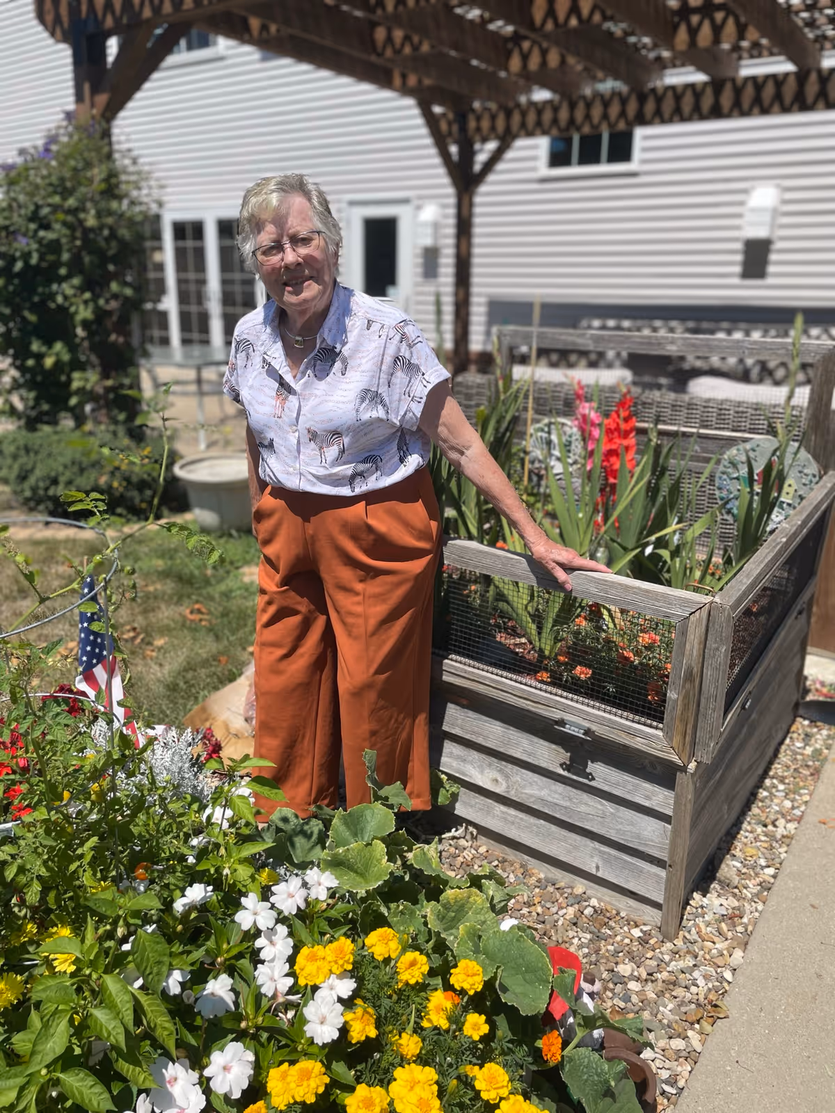 An elderly woman standing outdoors in a garden area next to a raised wooden planter box filled with flowers and plants. She is wearing a white shirt with a zebra print and orange pants, smiling at the camera. Behind her is a building with white siding and a wooden pergola.