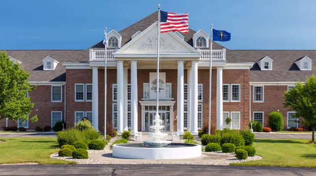 Front of a brick two-story building with white columns, flagpoles, and a fountain in a landscaped driveway.