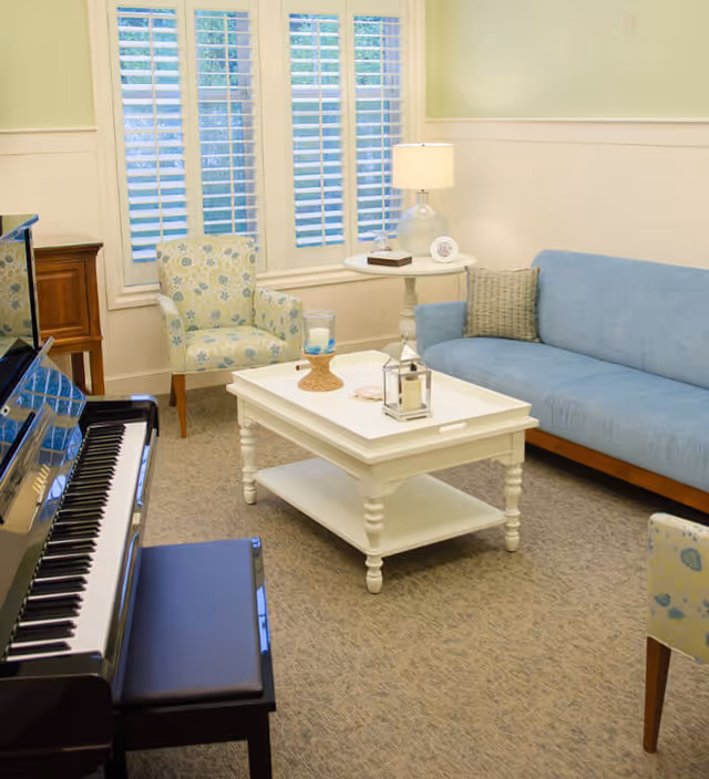 A cozy living room with a blue sofa, a floral patterned armchair, a white coffee table with decorative items, a side table with a lamp, and a black upright piano with a bench. The room has large windows with white shutters and light-colored walls.