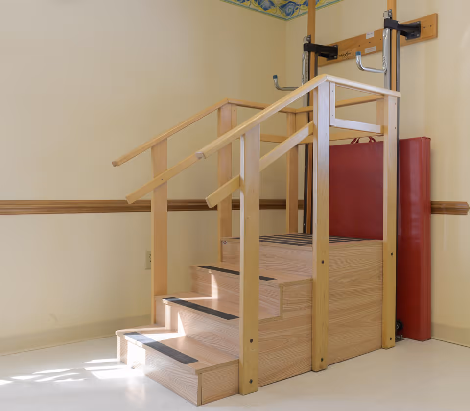 A wooden rehabilitation staircase with handrails on both sides and black anti-slip strips on each step, positioned indoors against a beige wall with a red padded mat behind it.