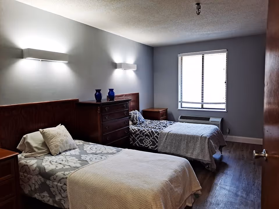 A bedroom with two single beds, each with patterned bedding and a pillow. Between the beds is a wooden dresser with two blue vases on top. There are two wall-mounted lights above the beds, a window with blinds, and a heating/cooling unit below the window. The room has wooden flooring and light gray walls.