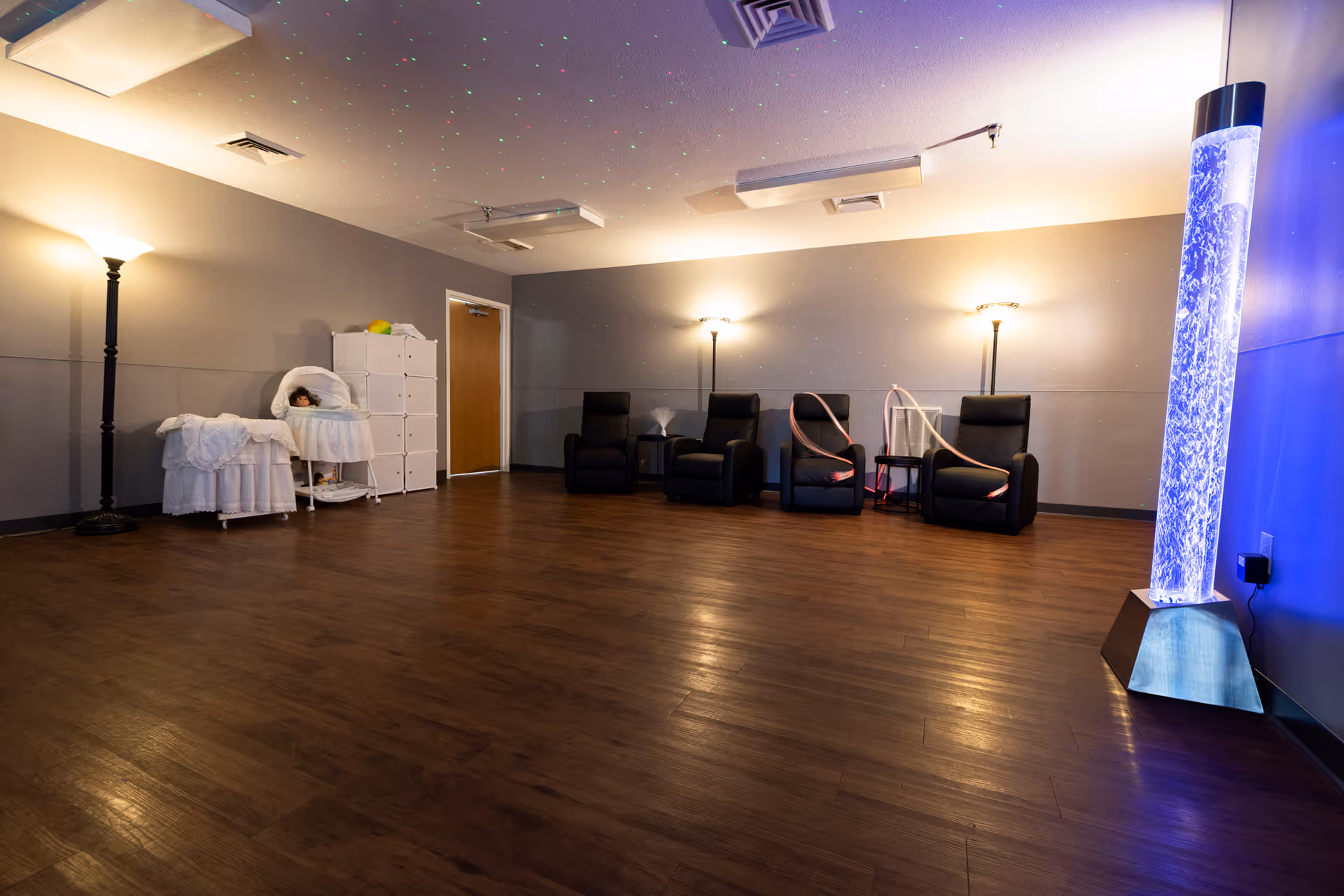 A spacious common room with reclining chairs, floor lamps, storage units and a tall illuminated bubble tube on a wood floor.