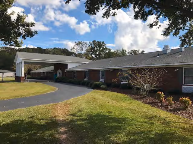 Exterior view of a single-story brick building with a covered entrance driveway, surrounded by grass and trees under a partly cloudy sky.