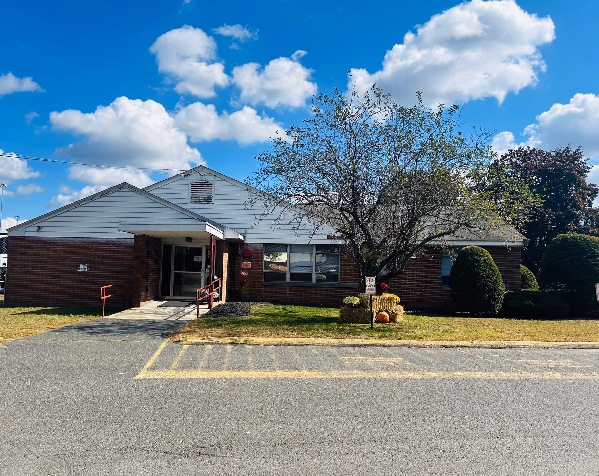 Exterior view of a single-story brick building with white siding on the upper part, a small covered entrance with red railings, a tree with some fall decorations including hay bales and a pumpkin, and a clear blue sky with scattered clouds.