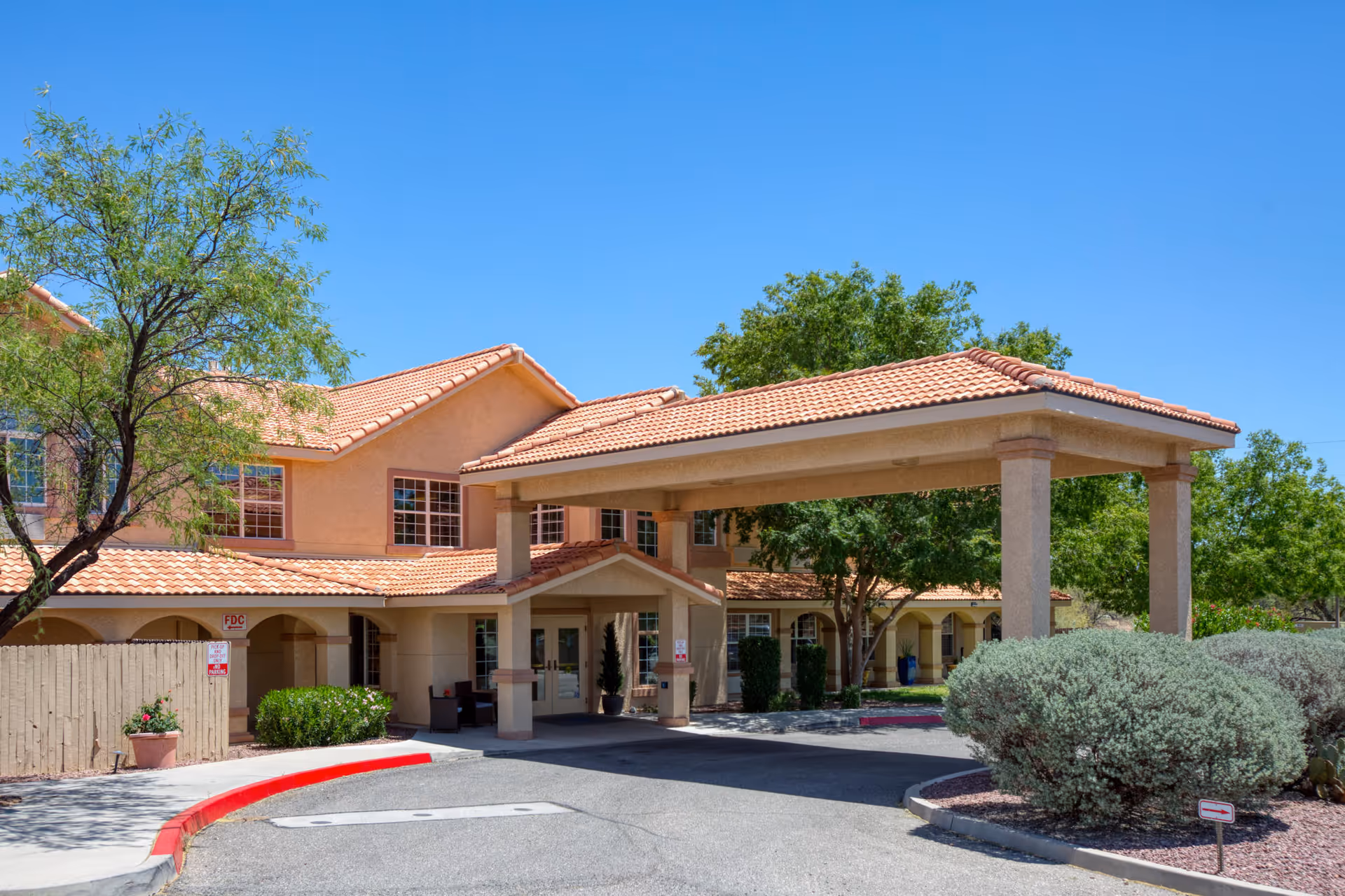 Exterior view of Vista Pointe at Green Valley senior living facility showing the entrance with a covered drop-off area, beige stucco walls, red tile roof, trees, and shrubs under a clear blue sky.