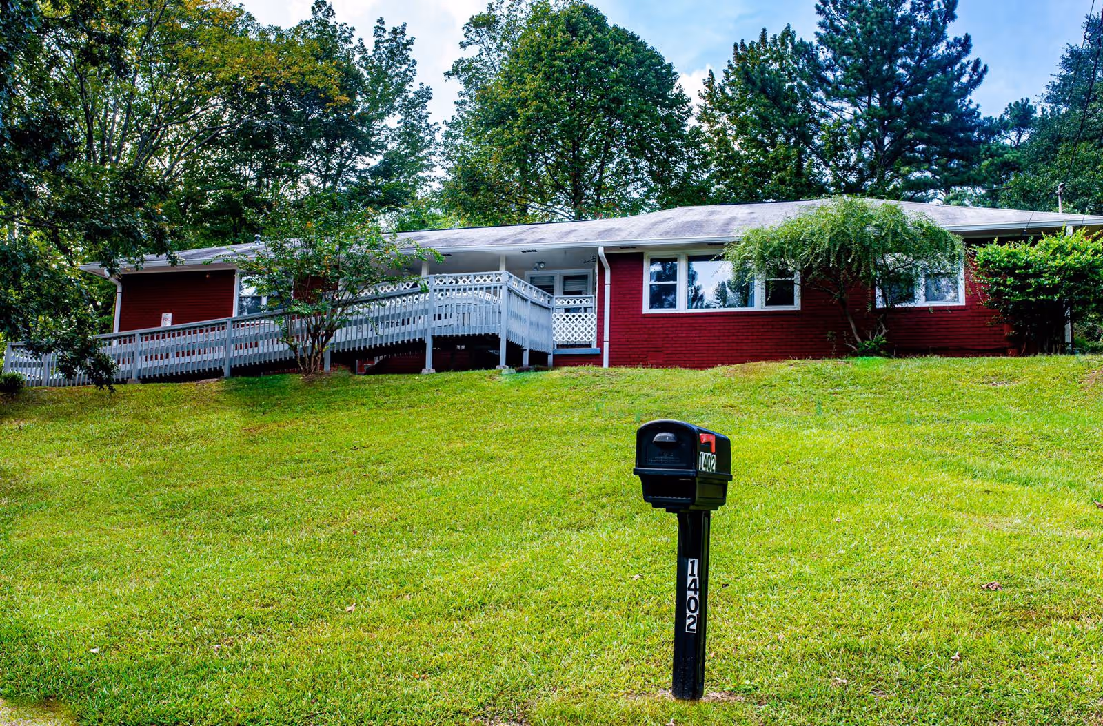 A single-story red brick building with white trim, surrounded by green trees and grass. There is a black mailbox with the number 1402 in the foreground, and a white wooden ramp leading to the entrance of the building.
