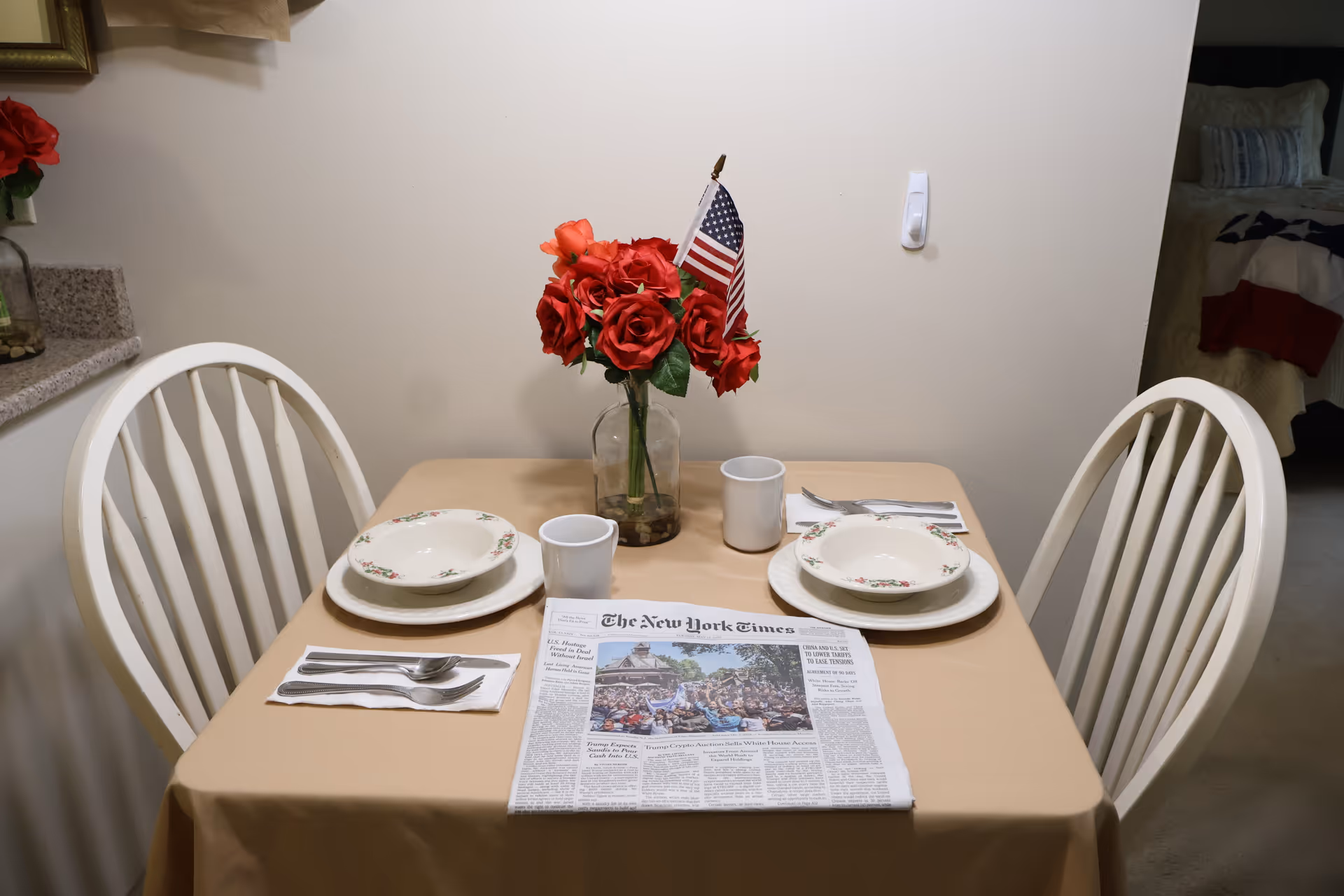 A small dining table set for two with white chairs, floral-patterned bowls and plates, white mugs, and silverware on napkins. A vase with red roses and a small American flag is in the center of the table. A folded newspaper is placed on the table. In the background, part of a bedroom is visible.