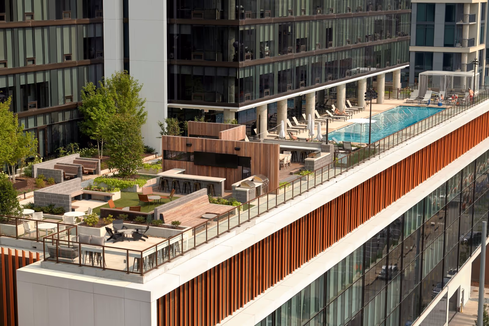 Rooftop outdoor area of a modern building featuring a swimming pool with lounge chairs, a seating area with tables and chairs, greenery including trees and plants, and a wooden structure with bar stools and grills.