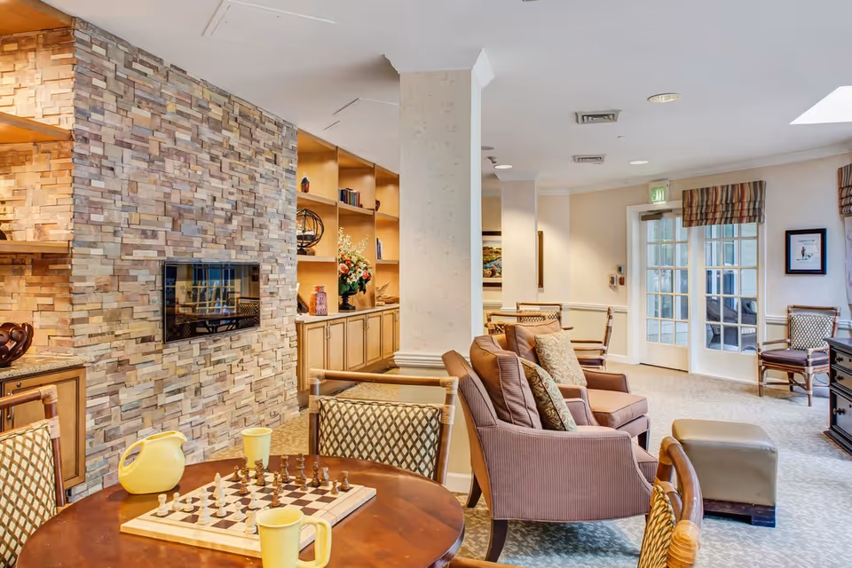 Bright communal lounge with upholstered seating, a stone-accent wall and mounted TV, and a table with a chessboard and mugs.