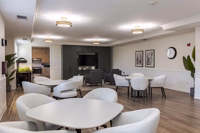 A spacious common area in a senior living facility with several round tables surrounded by white cushioned chairs. In the background, there is a dark green entertainment center with a flat-screen TV and two gray armchairs facing it. The room has wood flooring, white walls with wainscoting, two framed abstract artworks, a clock, and ceiling lights. A kitchen area is visible through an open doorway on the left side, along with some plants.