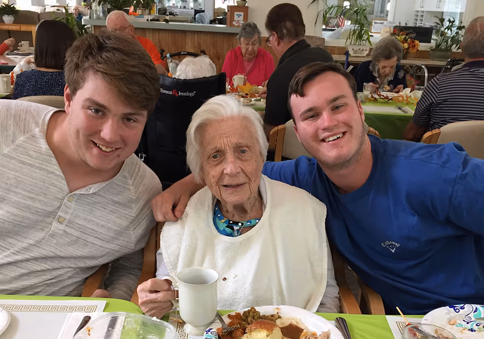 An elderly woman sits between two young men smiling at a dining table with plates of food in a communal dining room.