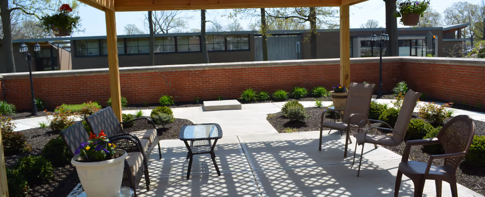 Outdoor patio area with a wooden pergola casting shadows on the concrete floor. There are several chairs and a small glass-top table arranged under the pergola. Surrounding the patio are landscaped garden beds with small bushes and flowers, bordered by a red brick wall. In the background, a building with large windows is visible.