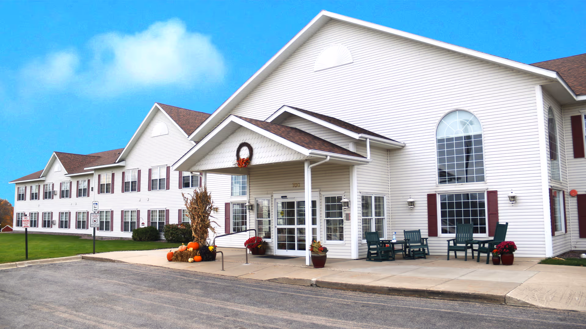 Front entrance of a two-story white senior living building with a covered entry, outdoor chairs, and seasonal pumpkin decorations.