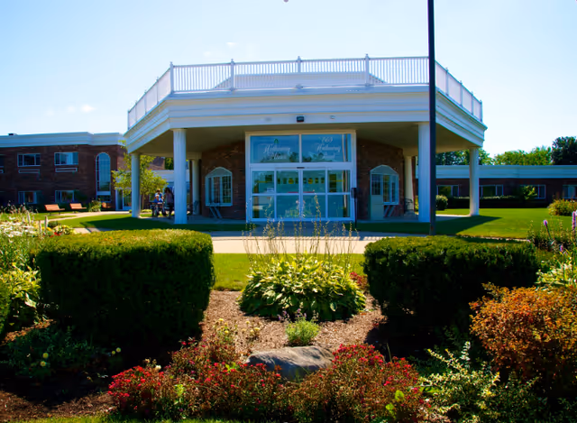 Front exterior view of Hathaway Manor Extended Care building with a covered entrance supported by white columns, surrounded by well-maintained bushes, flowers, and green lawn under a clear blue sky.