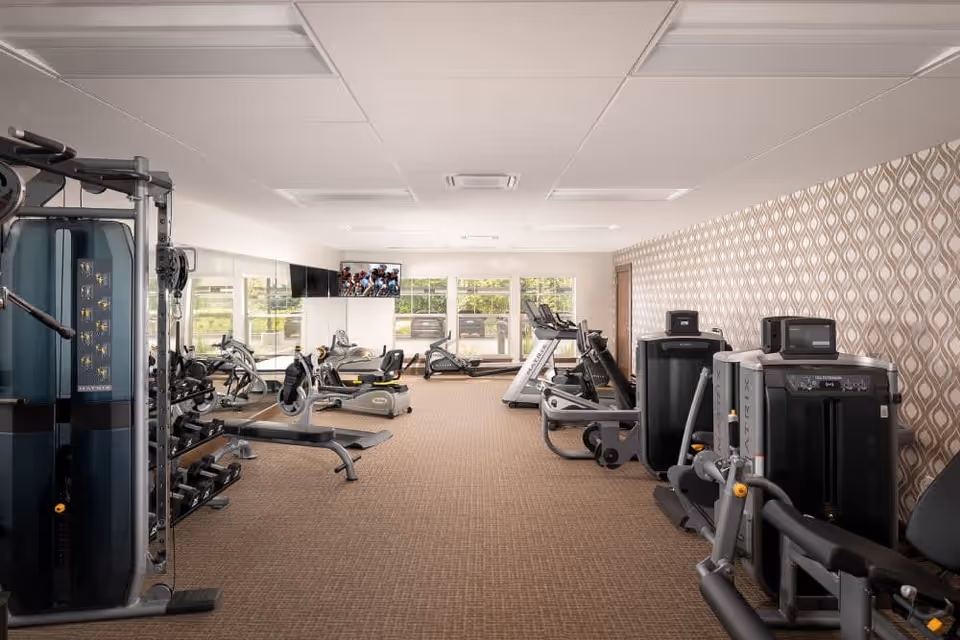 A well-lit fitness room with various exercise equipment including weight machines, stationary bikes, and treadmills. The room has large windows at the far end allowing natural light to enter, patterned wallpaper on one side, and a TV mounted on the wall.