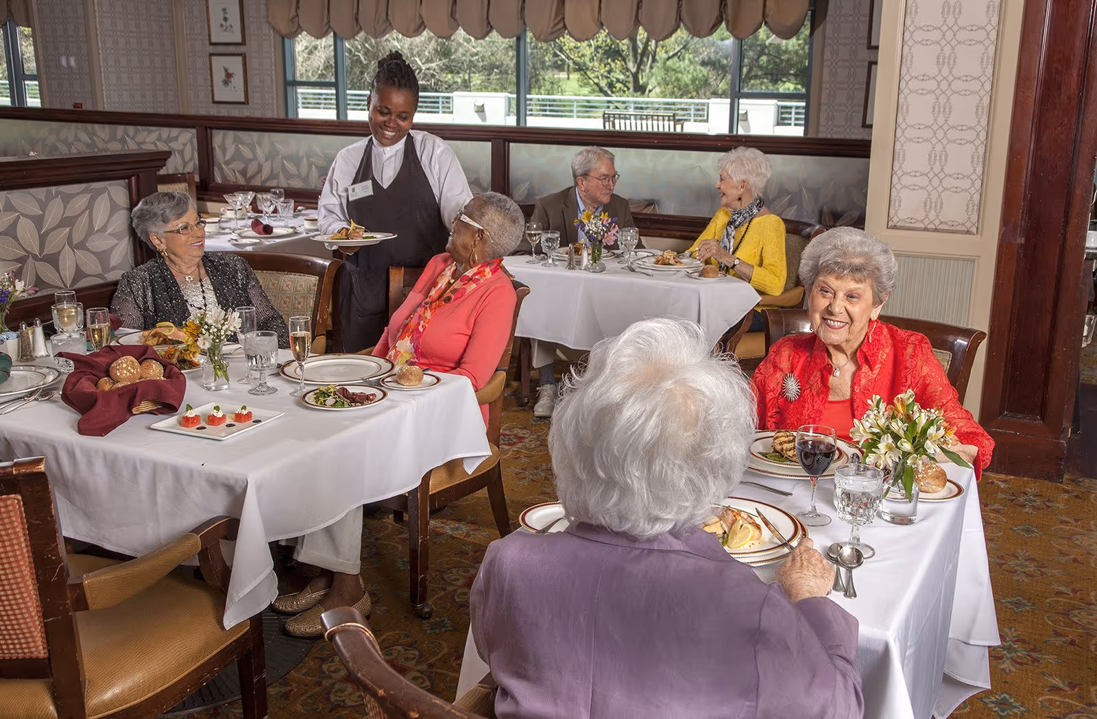 A dining room scene at Five Star Premier Residences of Chevy Chase with elderly residents seated at tables enjoying a meal. A smiling waitress is serving food to one of the tables. The room has large windows with a view of greenery outside, and the tables are set with white tablecloths, plates, glasses, and floral centerpieces.