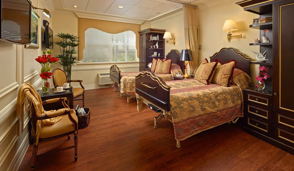 A well-decorated nursing home bedroom with two beds featuring ornate headboards and matching bedspreads. The room has wooden flooring, a window with blinds, two armchairs with a small table between them holding a vase of red flowers and tea set, and dark wood furniture including shelves and cabinets. The lighting includes wall-mounted lamps and a table lamp.