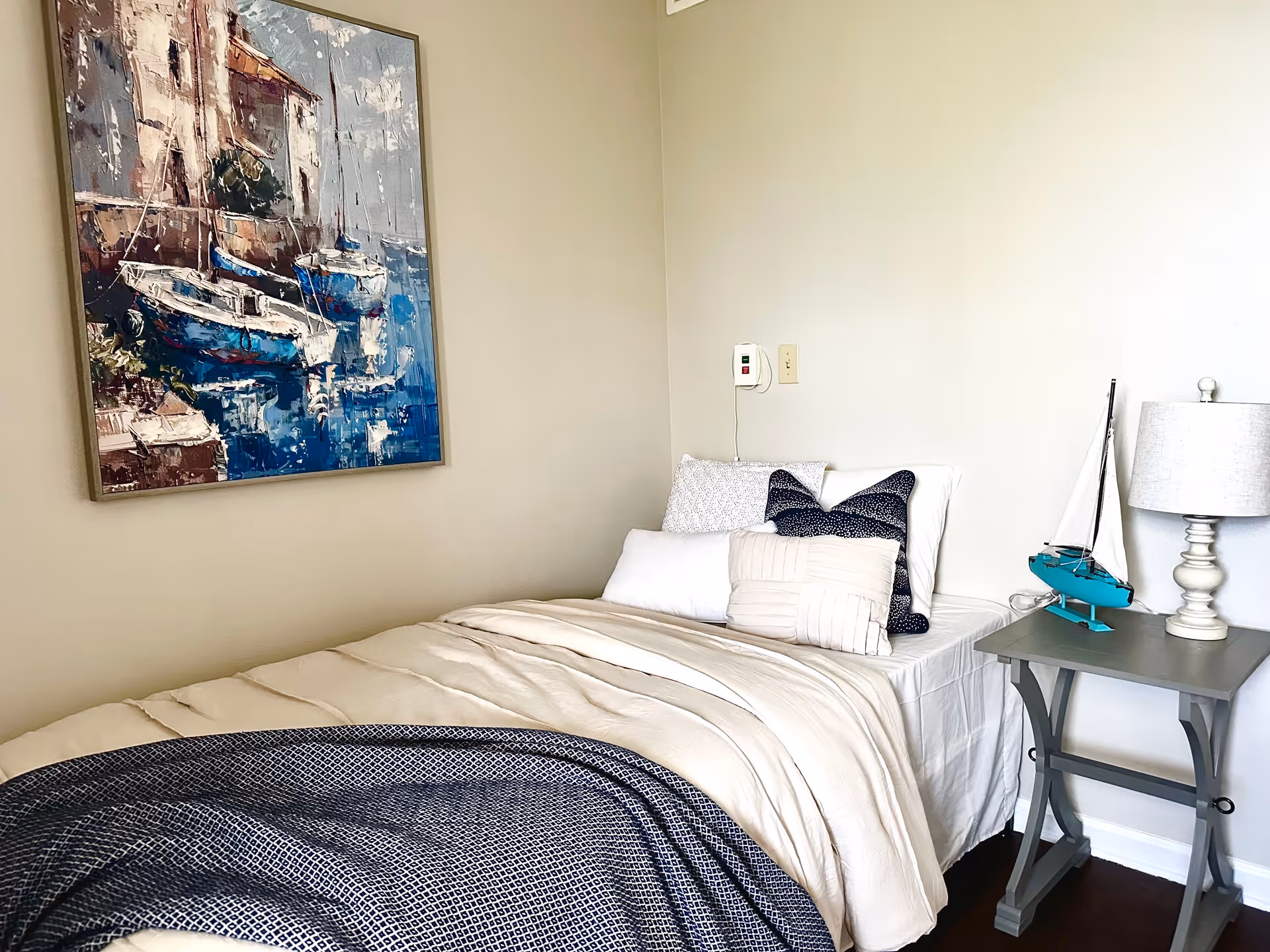 A neatly made single bed with white and navy blue pillows and a beige comforter in a room with light-colored walls. Next to the bed is a gray nightstand with a small decorative sailboat and a white table lamp. A framed painting of boats on water hangs on the wall above the bed.