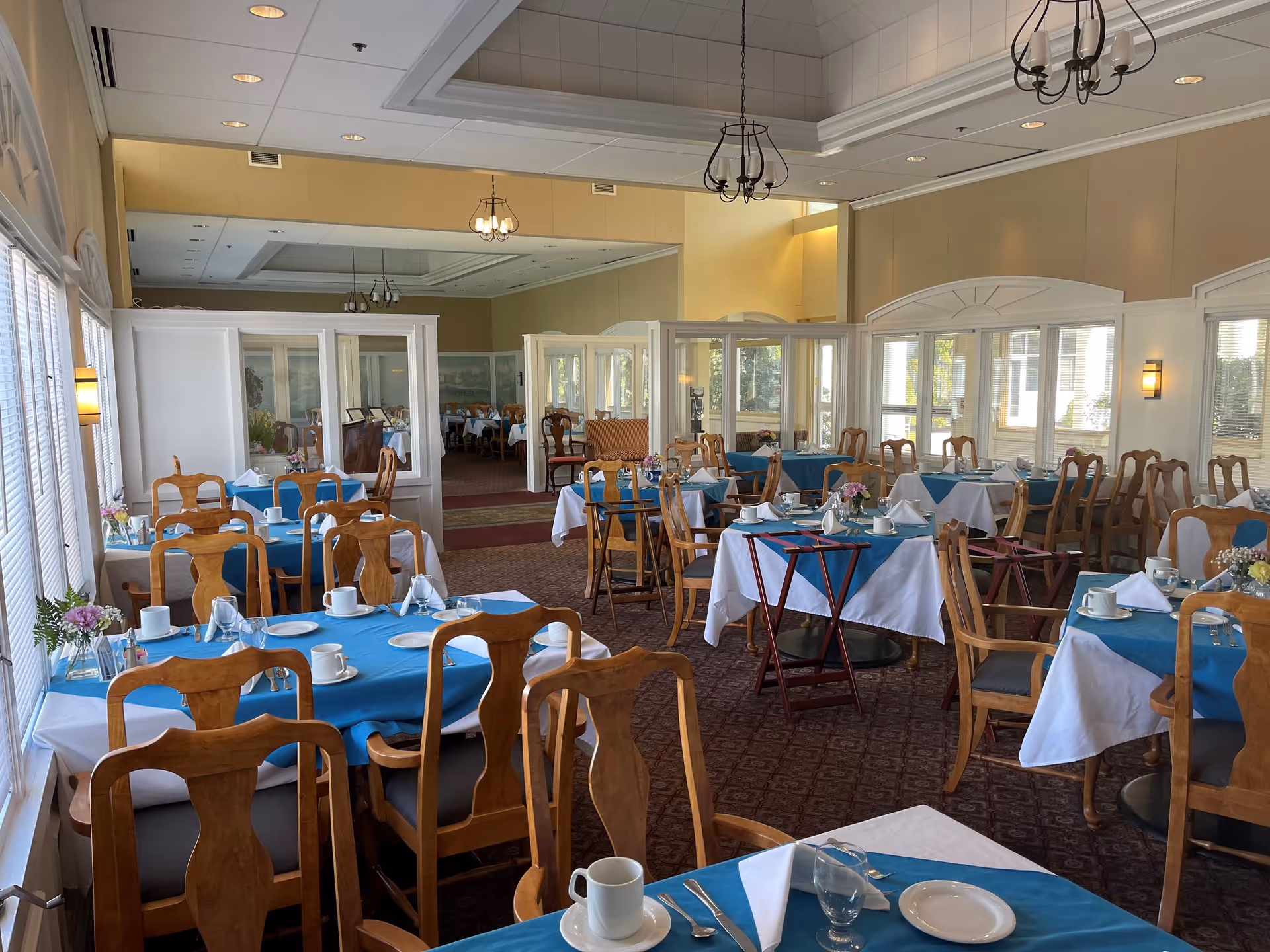 A bright dining room with multiple wooden tables and chairs arranged neatly. Tables are covered with white and blue tablecloths and set with white plates, cups, glasses, and silverware. There are small flower arrangements on each table. Large windows line the walls, allowing natural light to fill the room. The ceiling has recessed lighting and decorative chandeliers. A large mirror on one wall reflects the room, making it appear spacious.