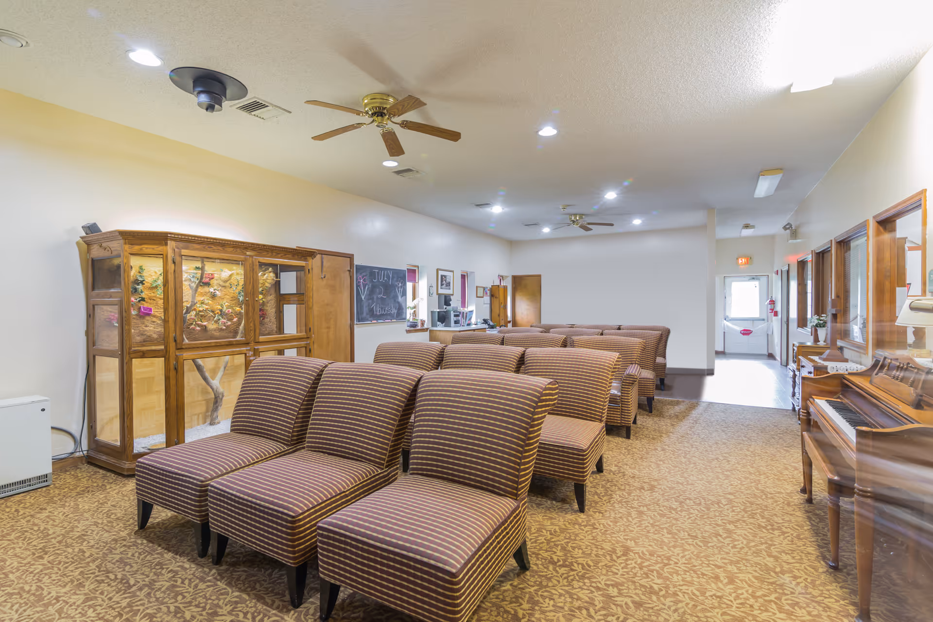 A senior living facility common room with several rows of striped upholstered chairs arranged facing forward. The room has a patterned carpet, a wooden piano on the right, a large wooden display cabinet on the left, and ceiling fans with lights. There is a chalkboard on the left wall with the date July 2 Thursday written on it, and a door at the far end of the room.
