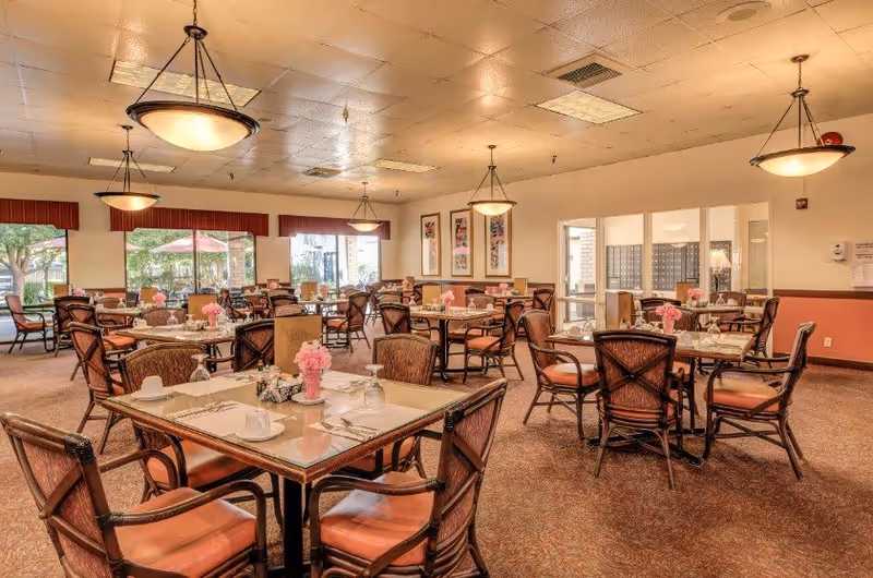 A spacious dining room with multiple tables and chairs arranged neatly. Each table is set with placemats, cups, utensils, and small flower vases with pink flowers. Large windows allow natural light to brighten the room, and there are hanging ceiling lights providing additional illumination. The walls are decorated with framed artwork and the room has a warm, inviting atmosphere.