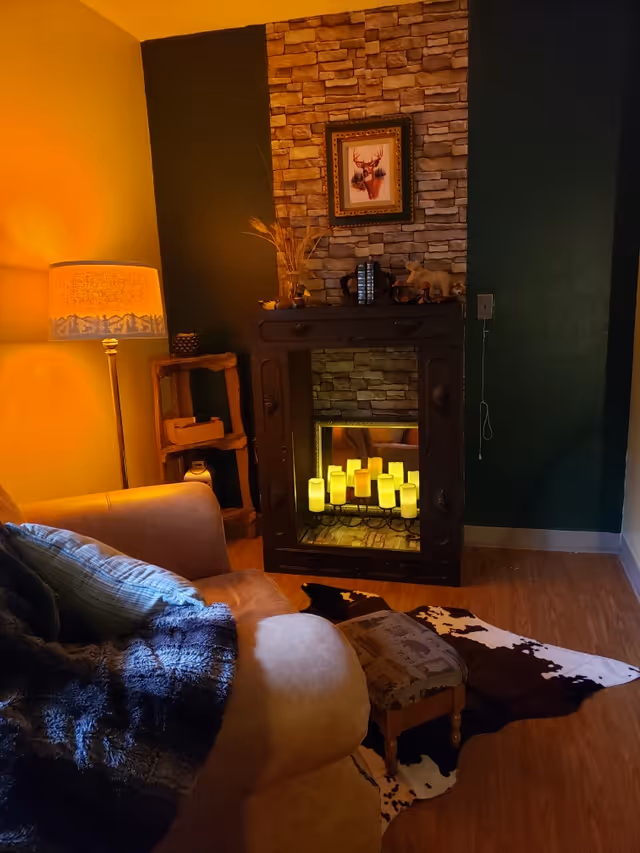 Cozy living room corner with a tan armchair draped with a dark blue blanket and a light blue pillow. Next to the chair is a small upholstered footstool on a cowhide rug. A floor lamp with a warm yellow shade illuminates the space. There is a decorative electric fireplace with multiple LED candles inside, set against a stone accent wall. Above the fireplace hangs a framed picture of a deer.