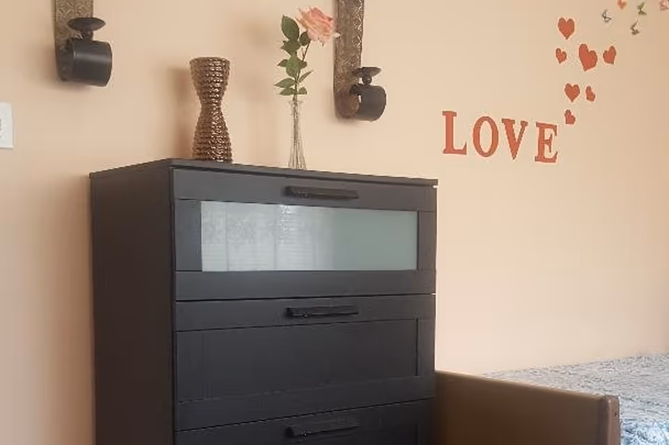 A black chest of drawers with three visible drawers, a decorative vase, and a single pink rose in a glass vase on top. The wall behind is beige with the word 'LOVE' and red heart-shaped decorations. Part of a bed with a blue patterned bedspread is visible on the right side.