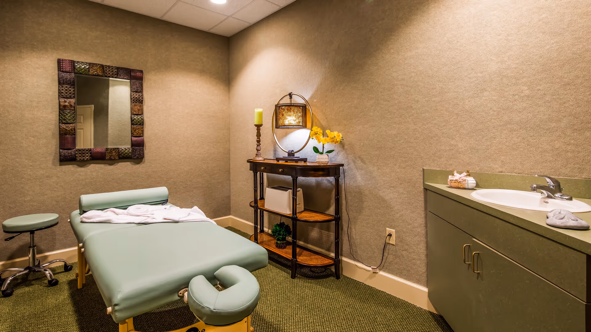 A small massage or therapy room with a green massage table covered partially with a white towel, a matching green stool on wheels, a decorative mirror on the wall, a small wooden shelf with a lamp, a candle, and a flower pot, and a green countertop with a sink and some folded towels.