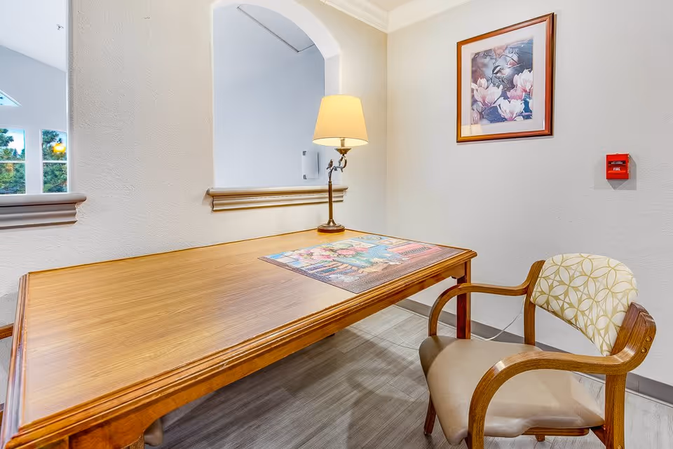 A wooden table with a decorative placemat and a lit table lamp on it, next to a cushioned chair with wooden armrests. The wall behind has a framed floral painting and a red fire alarm. There is an arched opening in the wall showing a glimpse of another room with windows.