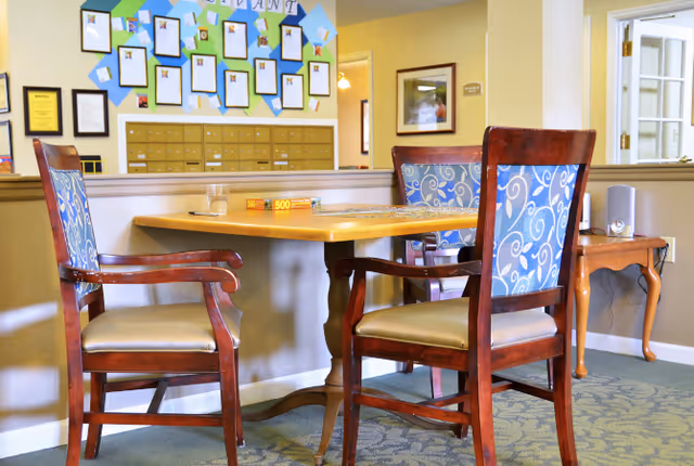 A small wooden table with two wooden chairs featuring blue patterned cushions in a well-lit room. On the table, there is a glass of water, a box of 500-piece puzzle, and a partially completed puzzle. The background shows a wall with multiple framed certificates and a set of mailboxes, along with a small wooden side table and a doorway with glass panels.