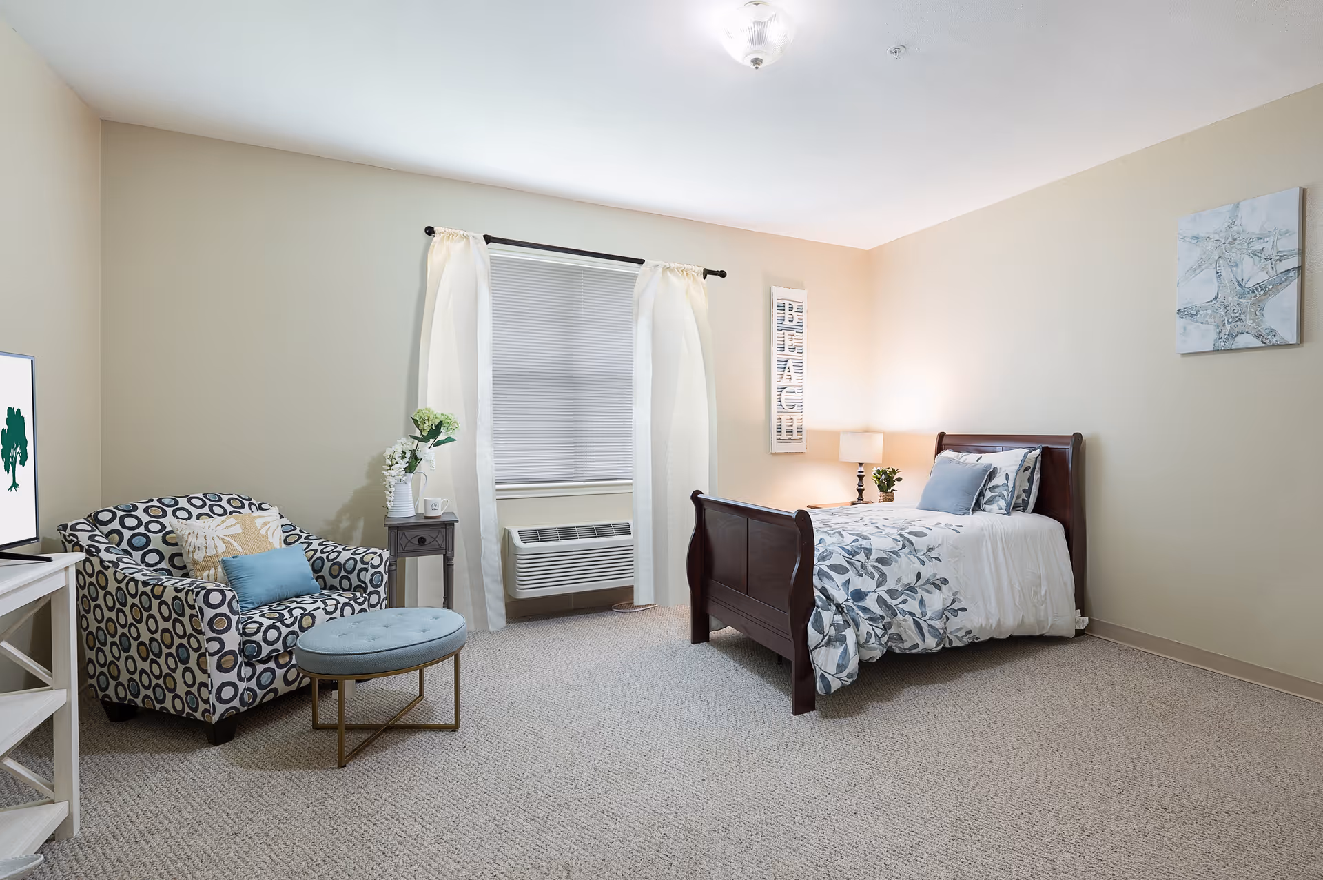 A cozy bedroom with a single wooden bed dressed in white and blue floral bedding, a patterned armchair with a blue cushion, a small round ottoman, a side table with a vase of flowers, a window with white curtains and blinds, a wall decoration with the word 'BEACH', and a starfish artwork on the wall.