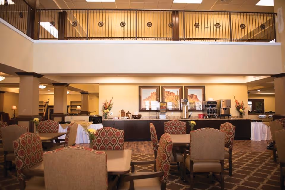 A spacious dining area in a senior living facility with multiple tables and cushioned chairs arranged neatly. The room features warm lighting, patterned carpet, and a counter against the far wall with coffee and beverage dispensers, decorative flowers, and framed landscape artwork above it. The upper level has a railing overlooking the dining space.