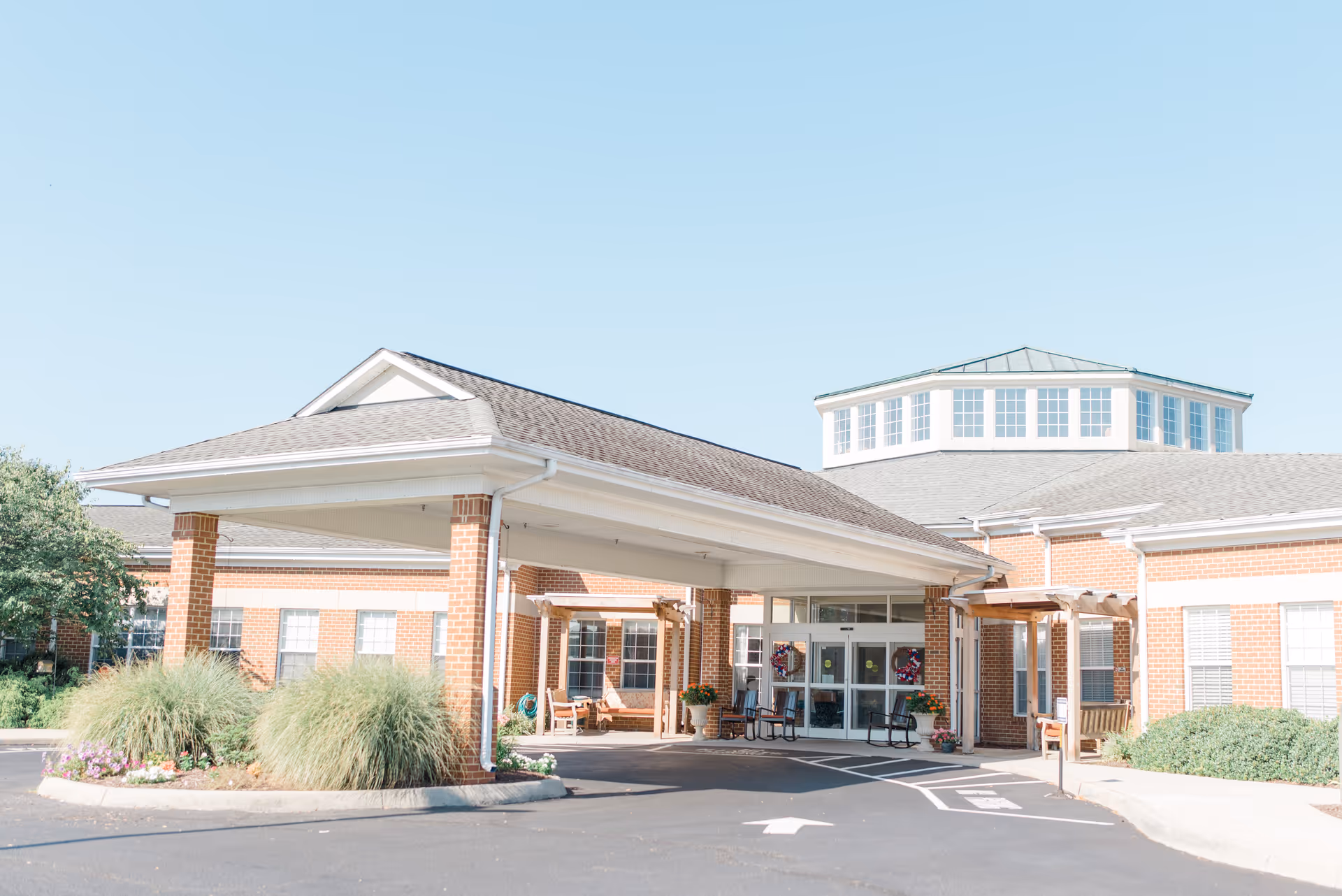 Covered entrance and porte-cochere of a brick assisted living building with glass double doors, benches, and landscaping.