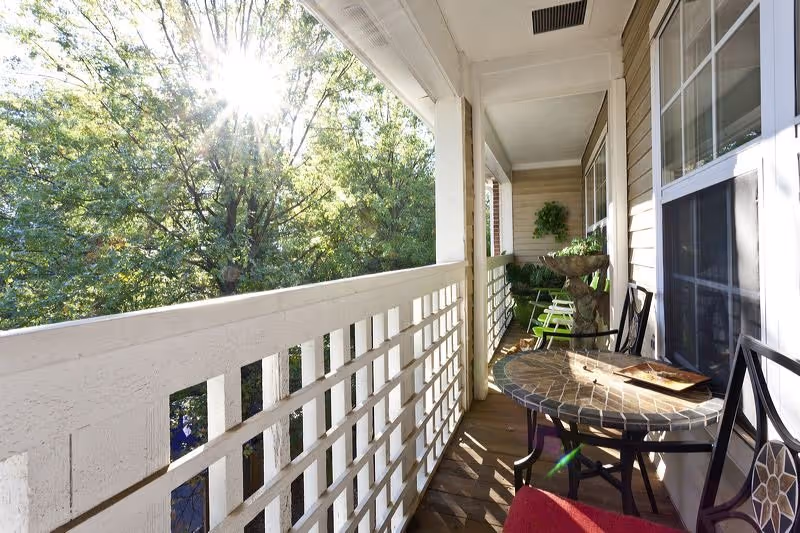 A sunlit balcony with a tiled round table and chairs, overlooking green leafy trees. The balcony has a white railing and beige siding on the building wall, with potted plants and additional seating further down the balcony.