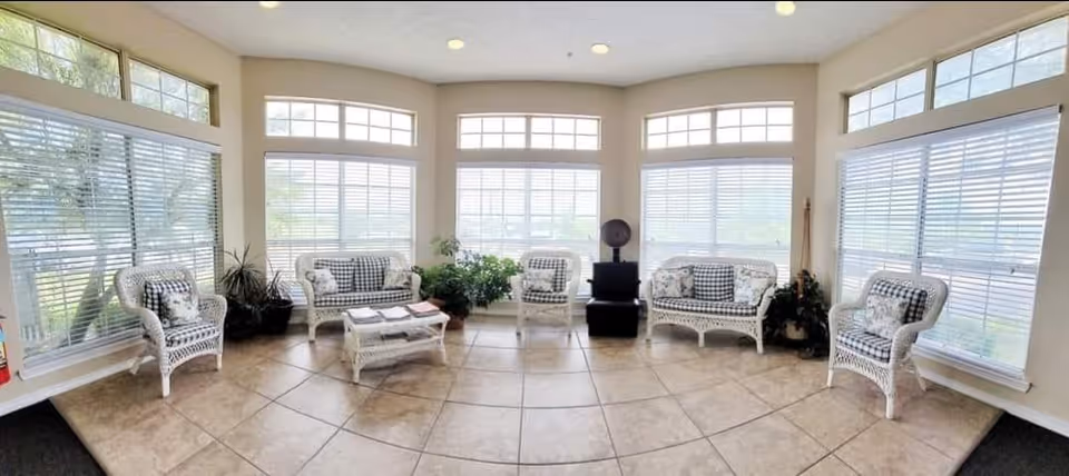 A bright and spacious living room area with large windows featuring white blinds, allowing natural light to fill the space. The room is furnished with white wicker chairs and loveseats with black and white checkered cushions, a matching wicker coffee table with magazines on top, and several green potted plants placed near the windows. The floor is tiled with large beige tiles, and the walls are painted a light color.