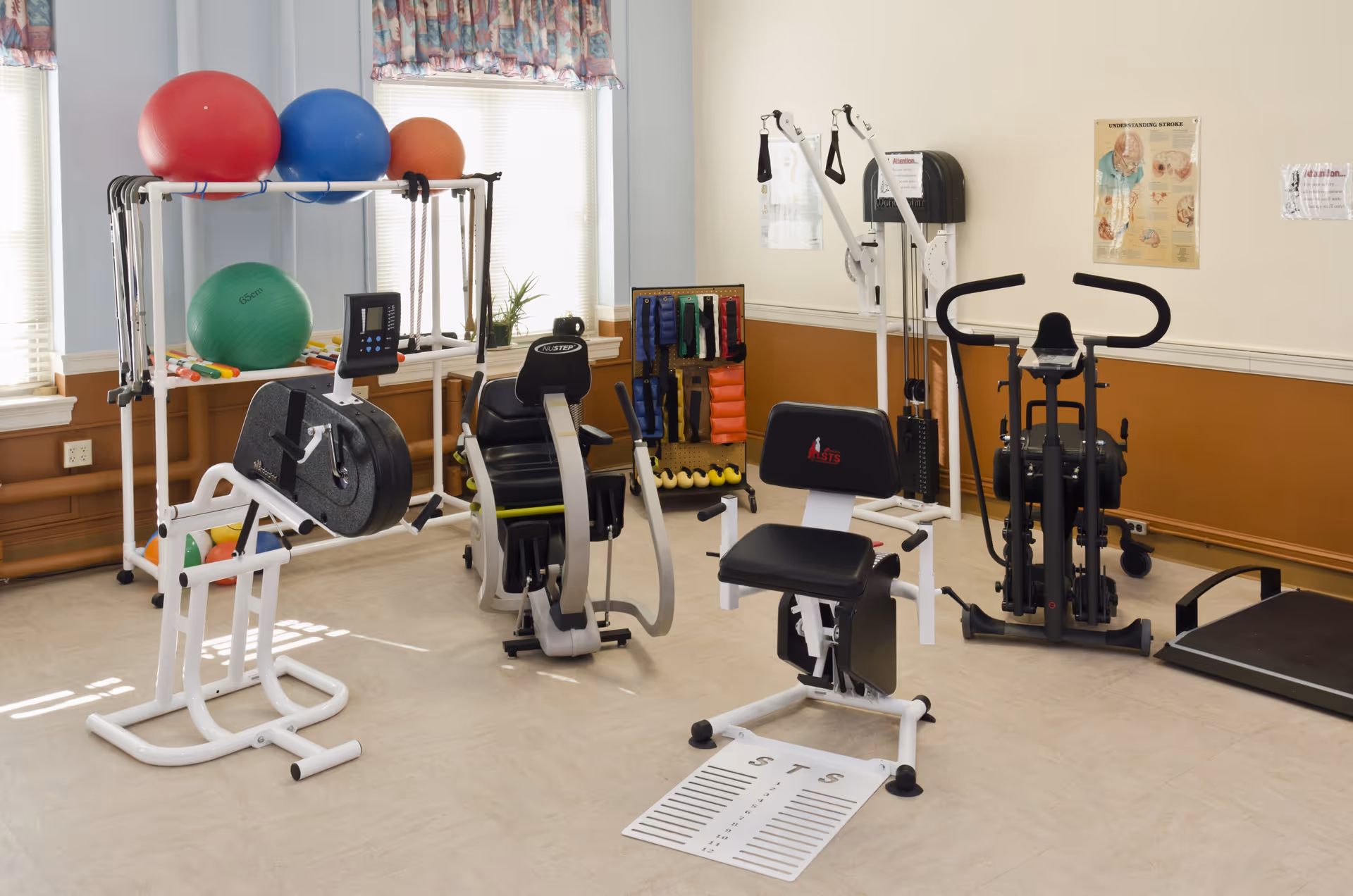 A small exercise room with various fitness equipment including stationary bikes, resistance bands, exercise balls, and a treadmill. The room has light-colored walls with a brown lower half and windows with floral curtains letting in natural light.