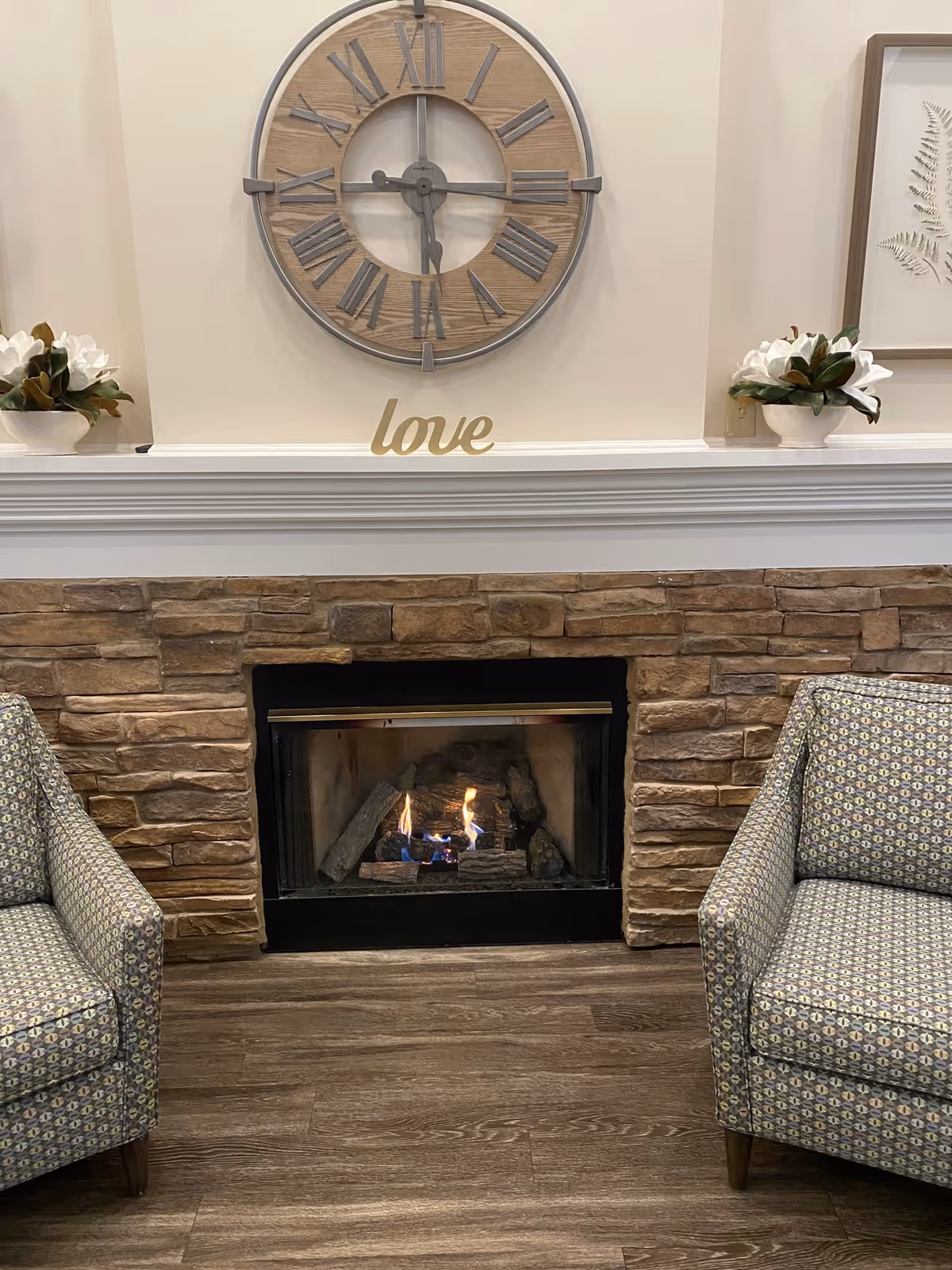 A cozy indoor seating area featuring a stone fireplace with a small fire burning. Above the fireplace is a large wall clock with Roman numerals and a decorative word 'love' placed on the mantel. On either side of the fireplace are patterned armchairs and white flower arrangements in white pots.